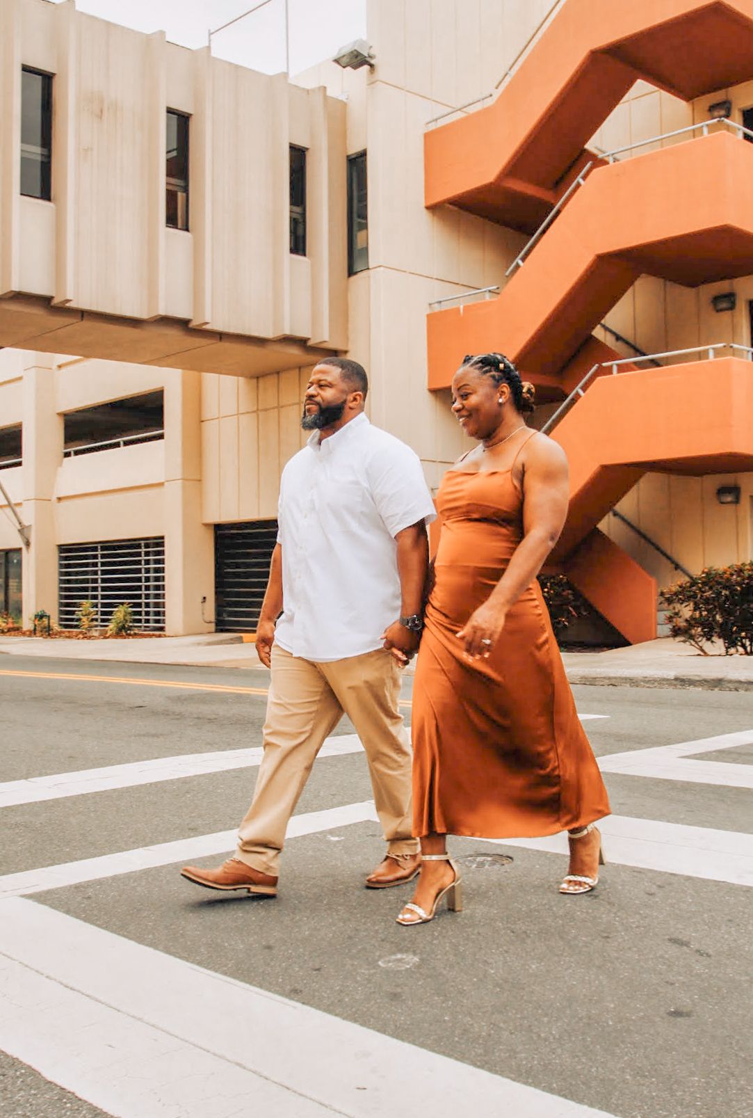 A couple walks together on a crosswalk with orange stairs and modern architecture in the background.