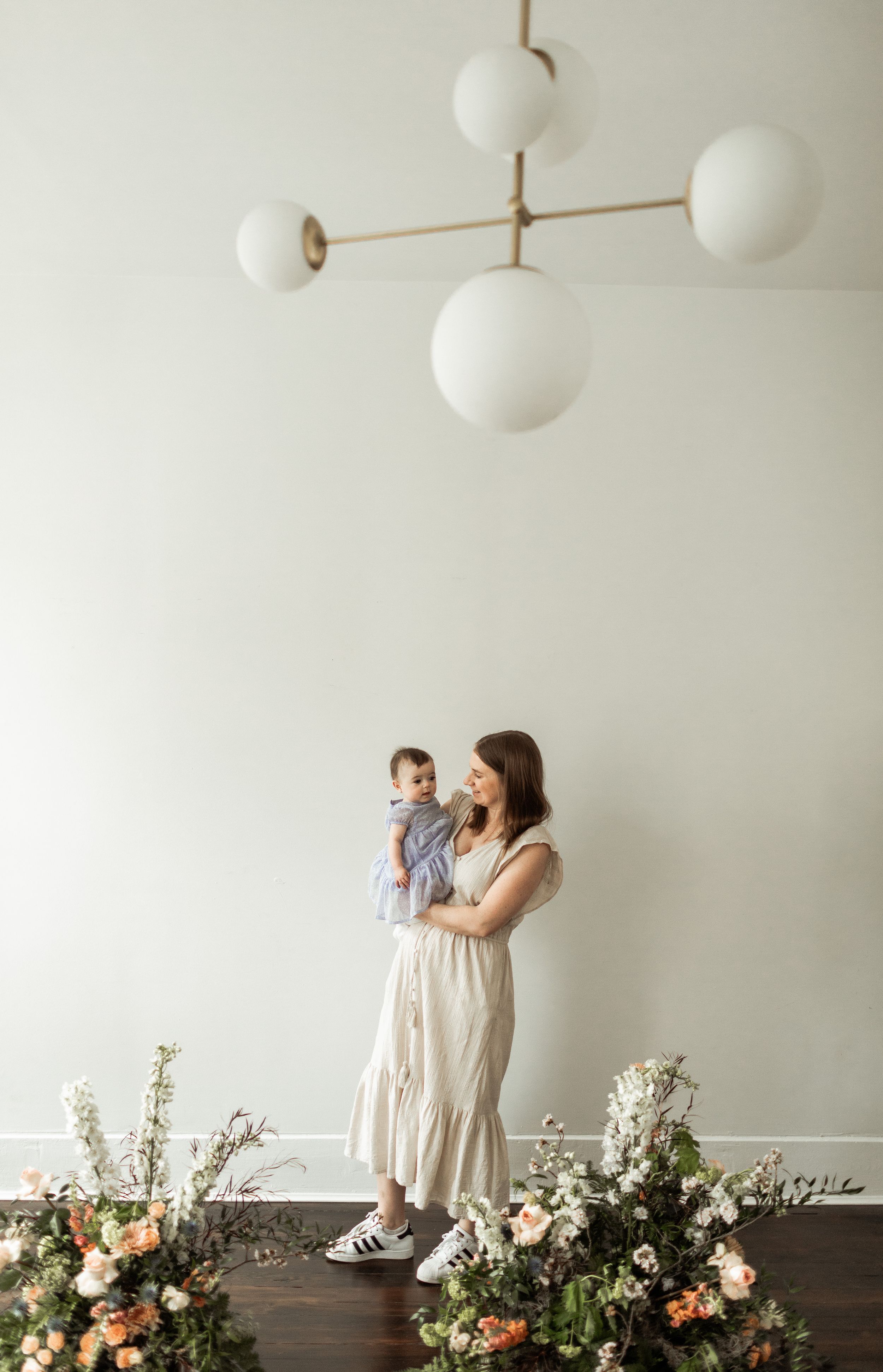 Modern white globe pendant light hangs above a mother and child standing among floral arrangements on dark wood floor.