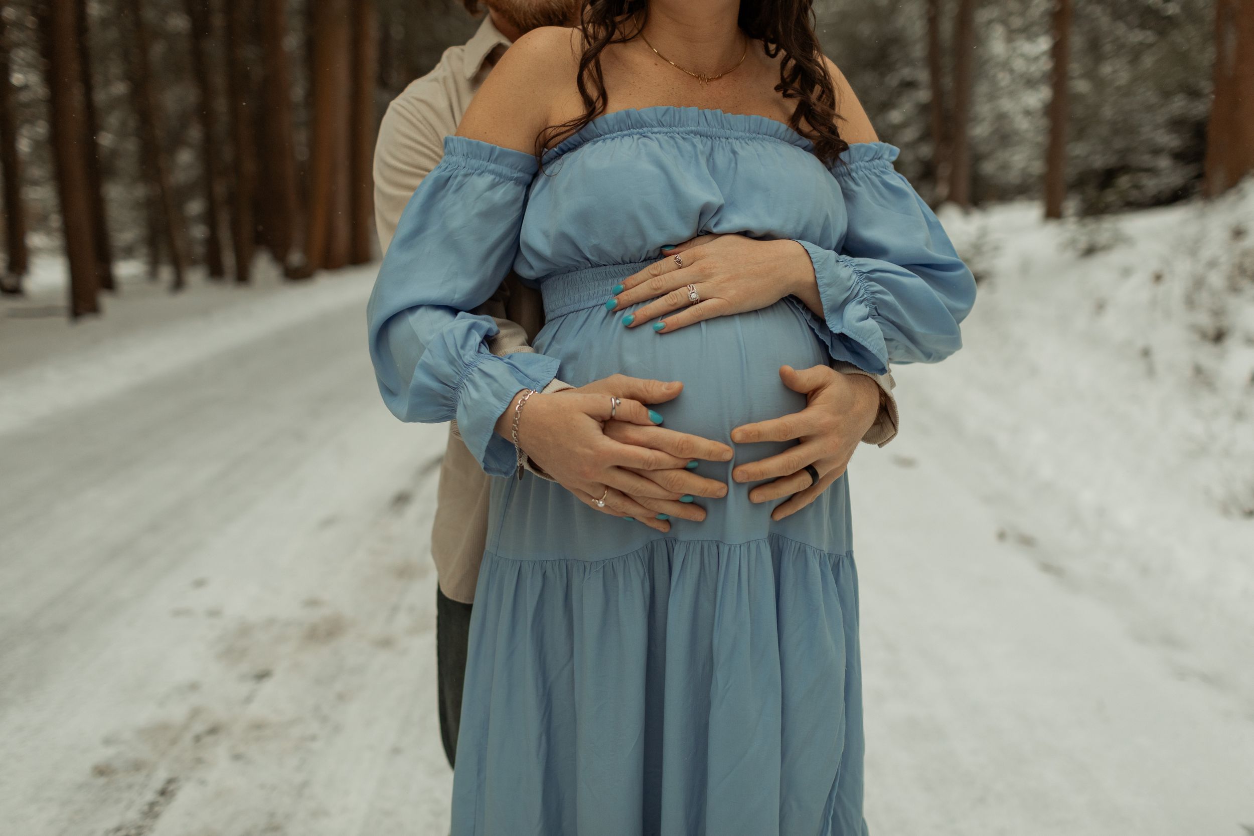 Expectant mother in light blue off-shoulder dress cradles her baby bump while standing on snowy forest path.