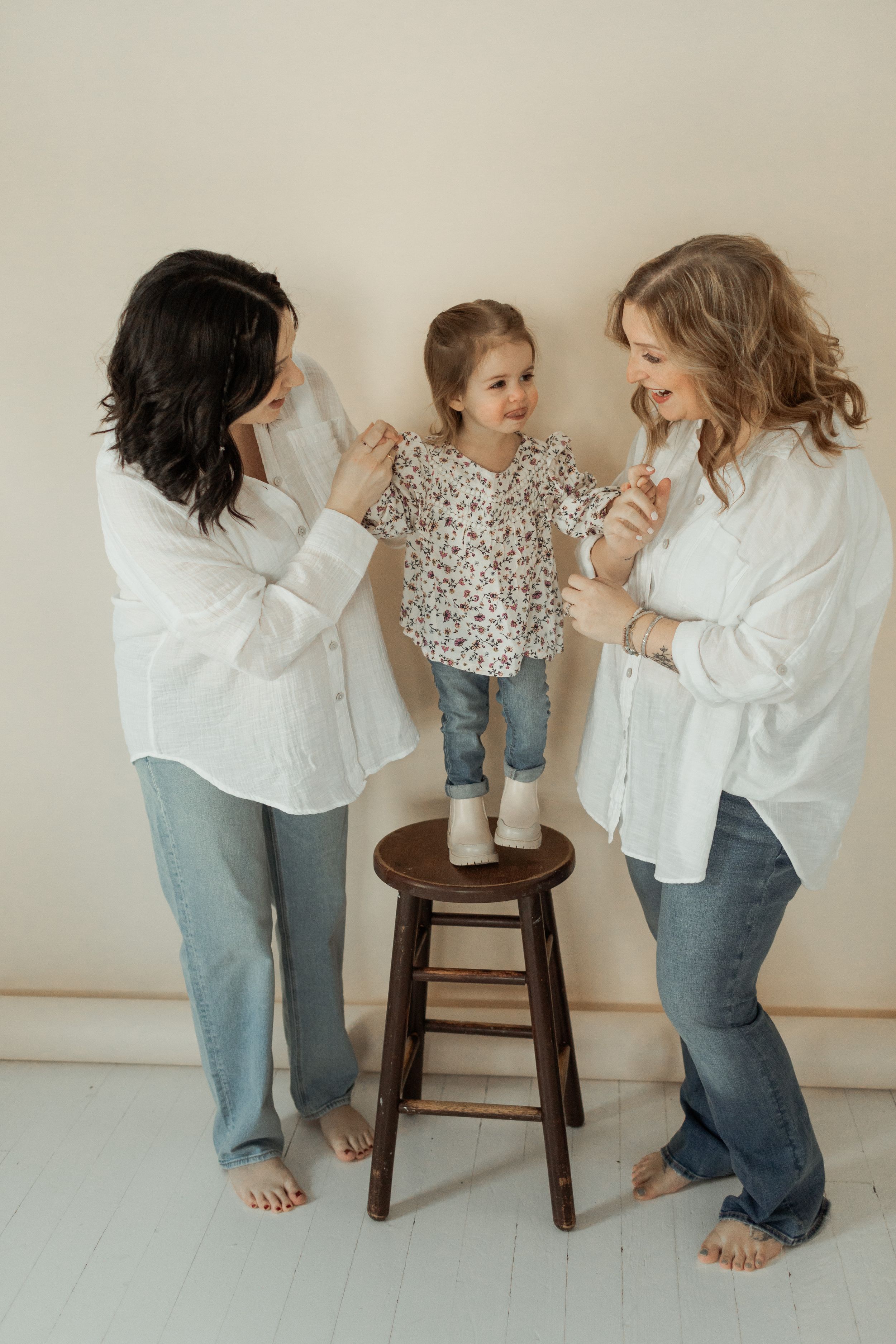Three people in casual outfits interact playfully around a wooden stool, creating a lighthearted moment in a bright room.