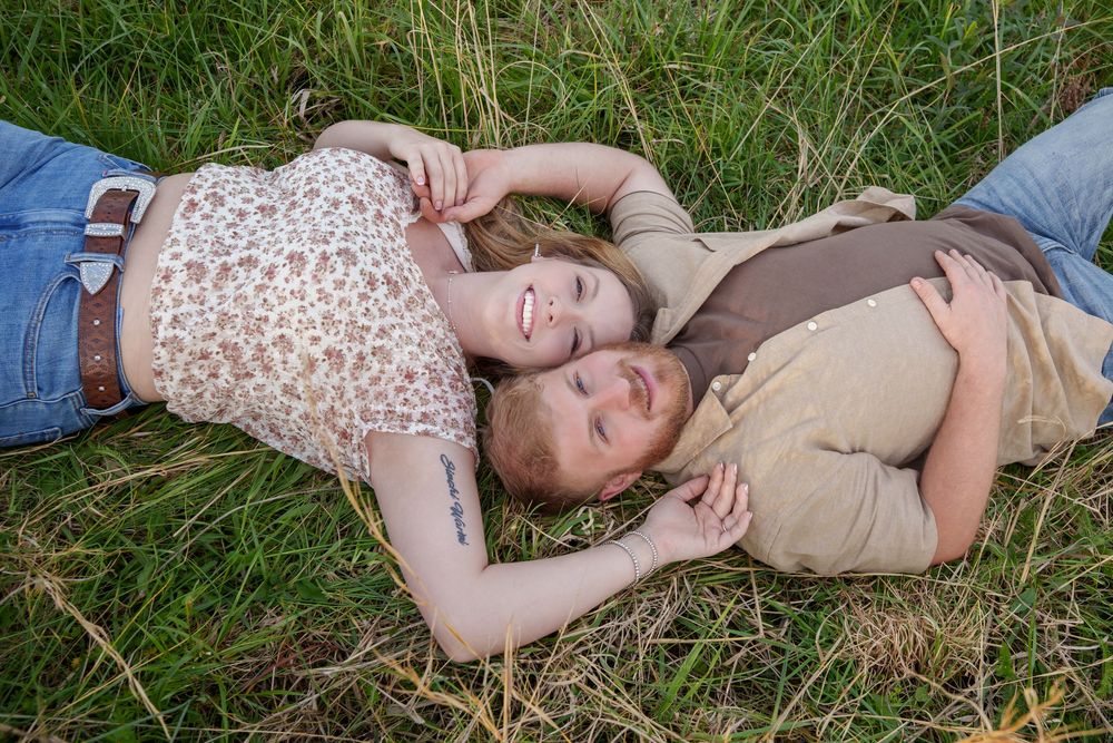 Two young people lying in green grass, smiling and holding hands in a romantic outdoor portrait.