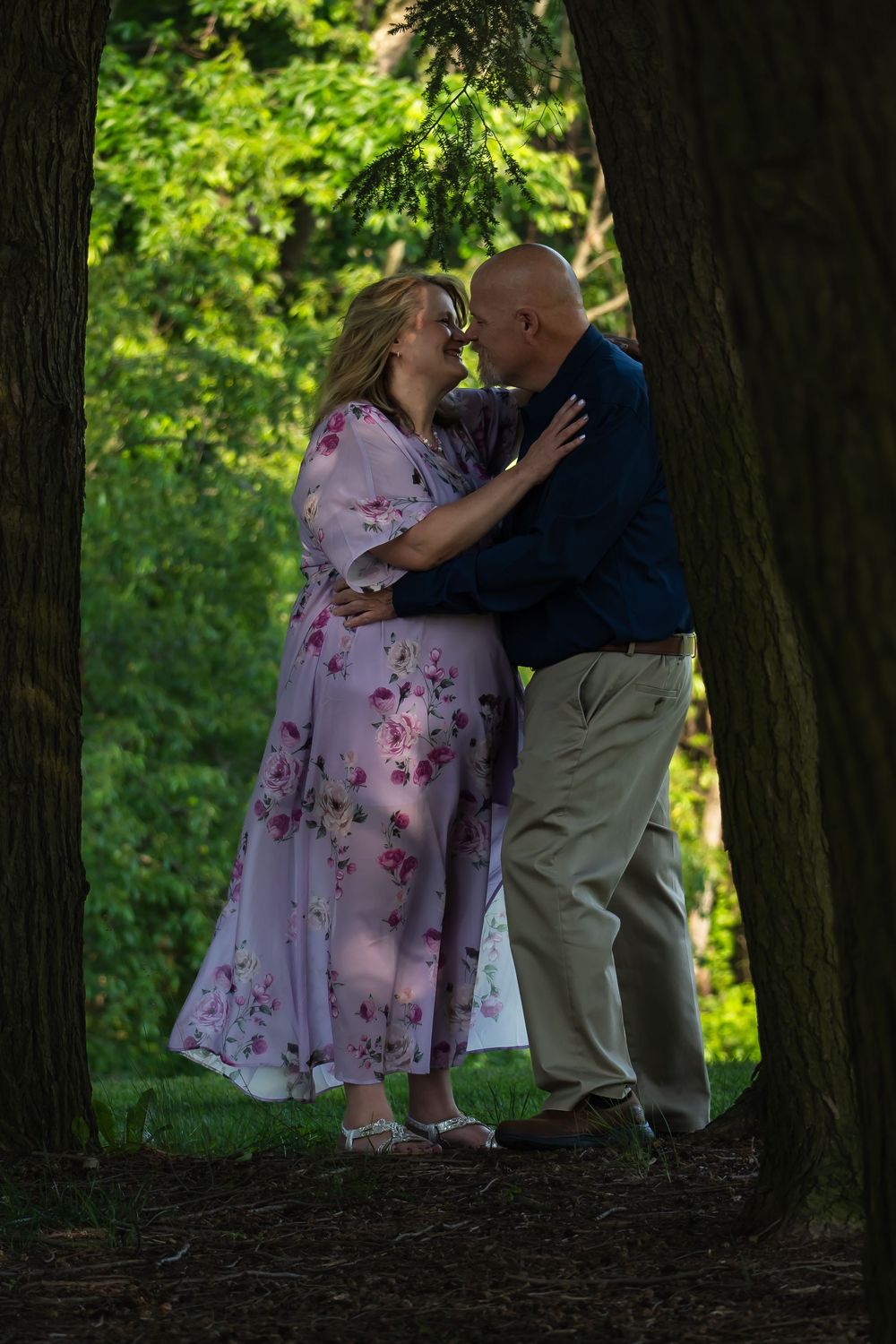 A romantic couple embraces between trees in a woodland setting, with her wearing a pink floral dress.