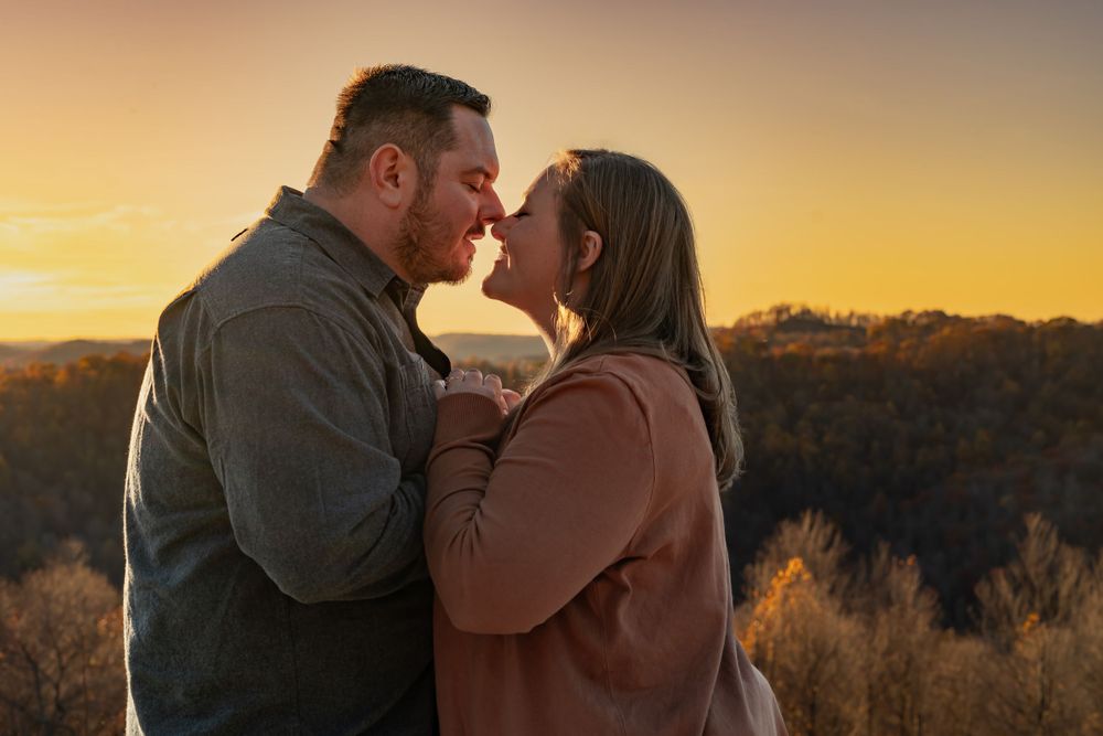 Intimate couple embracing during sunset with warm orange sky and trees in background.