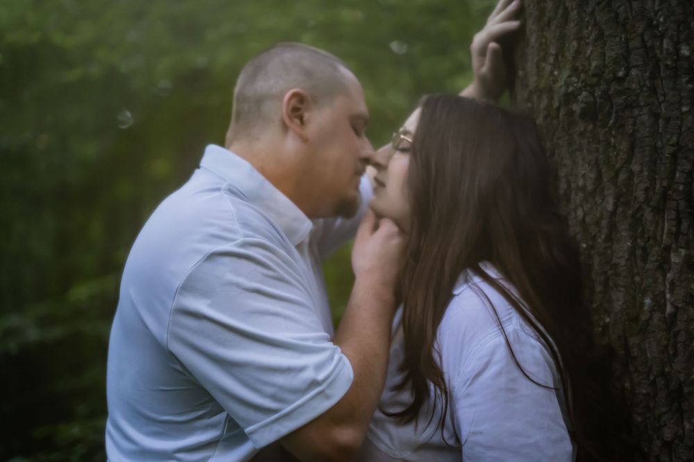 A tender moment shared between a couple by a tree trunk in a wooded area.