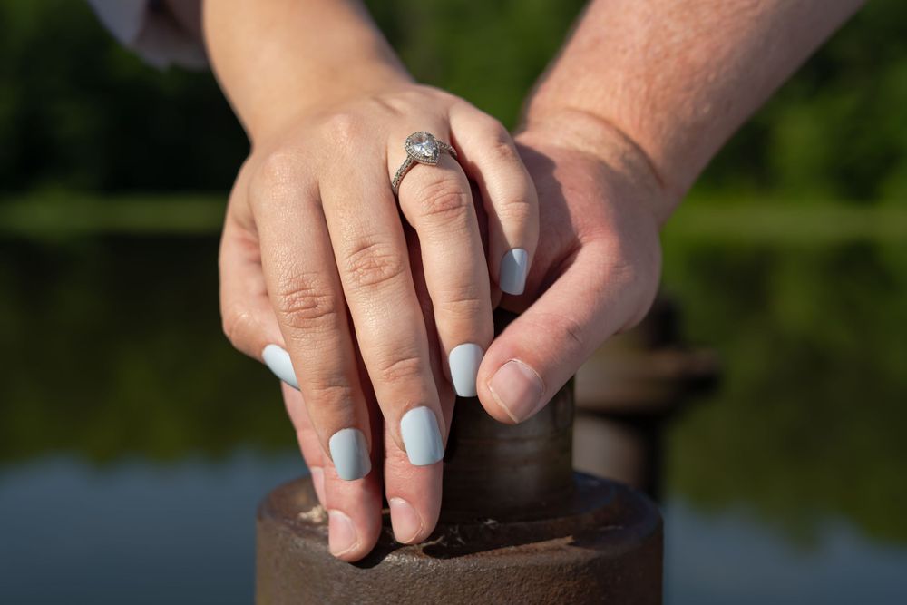 Close-up of hands with engagement ring resting on a wooden surface near water.