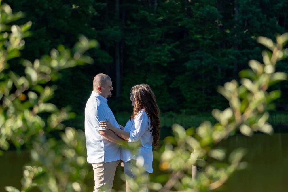 A romantic couple embraces by a serene lake surrounded by green foliage on a sunny day.