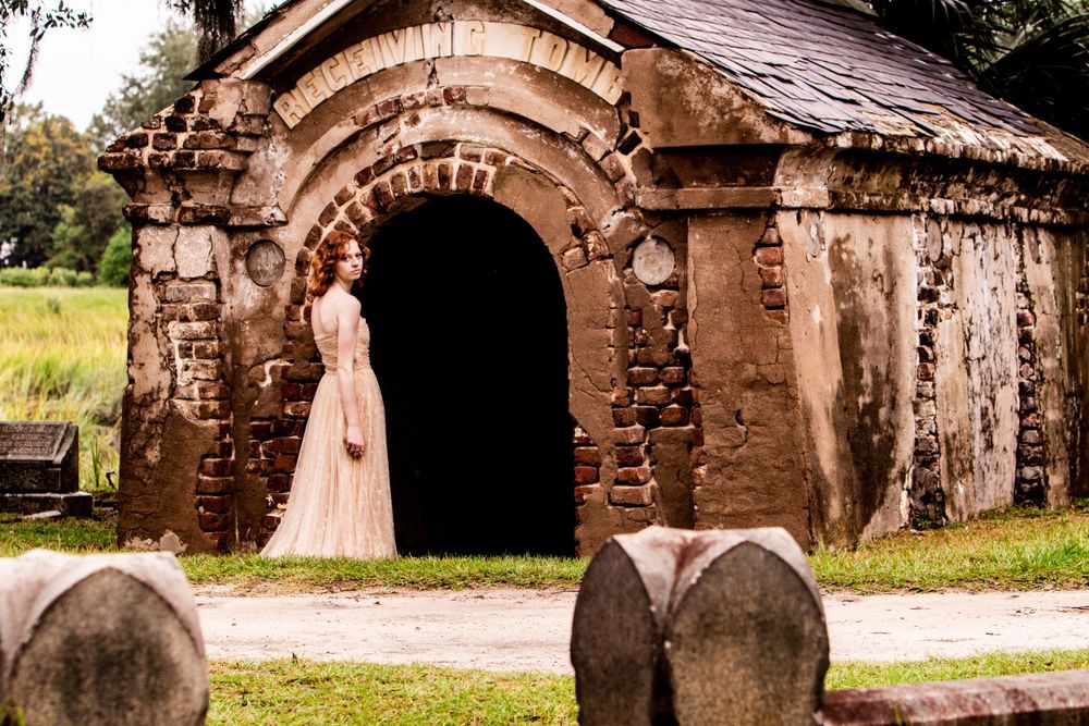 An elegant cream-colored vintage dress displayed at the entrance of a rustic stone chapel with gravestones in the foreground.