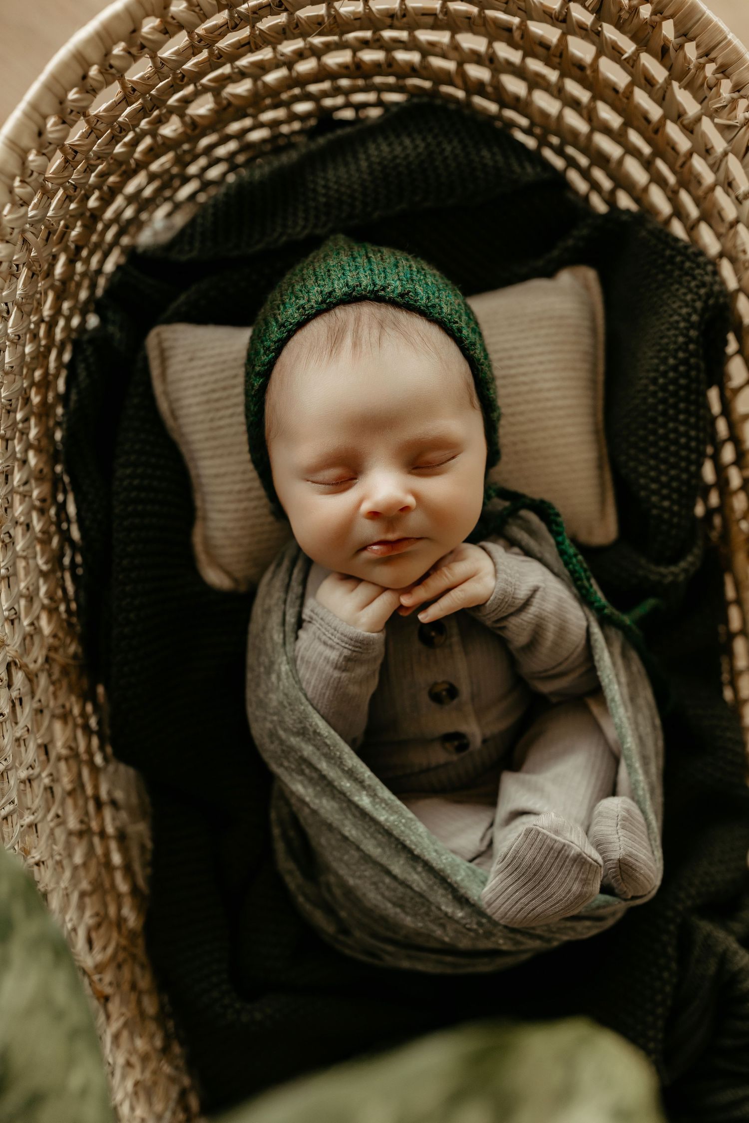 A sleeping baby photographed from above in a woven basket wearing a dark green knit cap and gray sweater.
