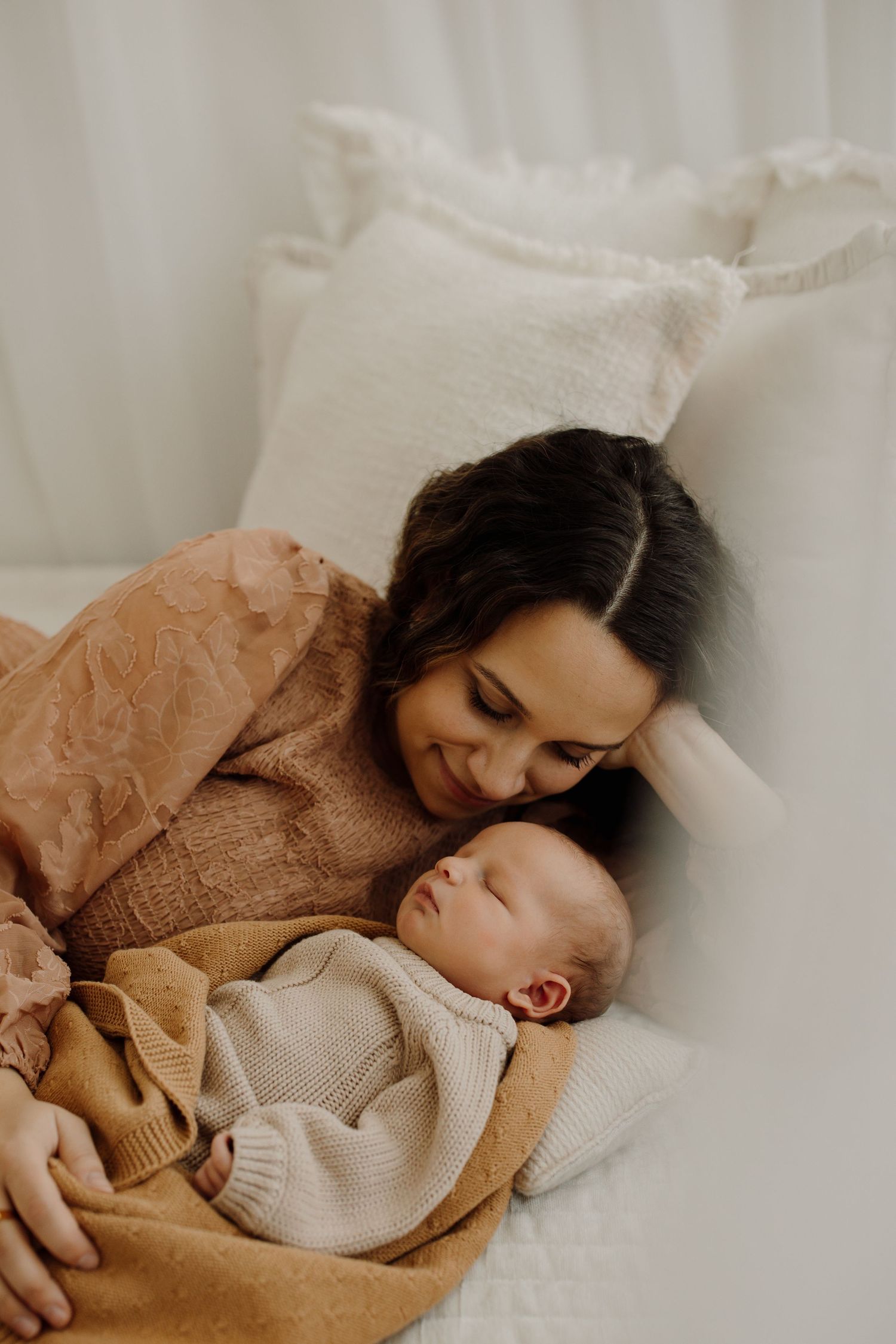 A tender moment of mother and baby snuggling together on white bedding in warm earth tones.