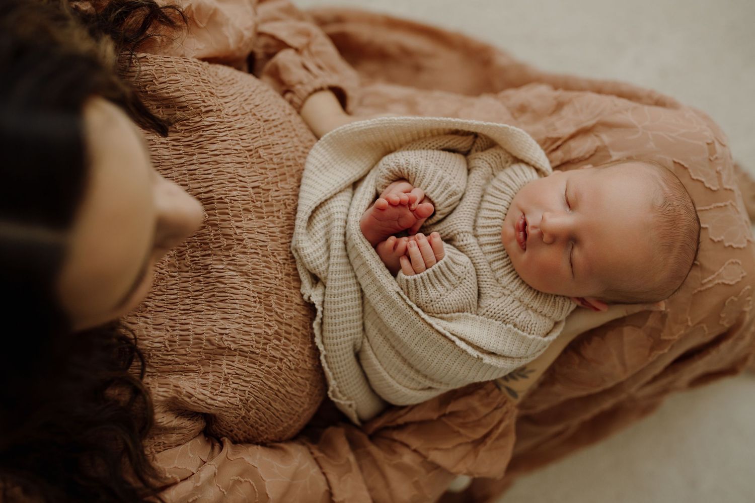 Baby wearing cozy knit sweater lies on soft brown blanket in warm natural lighting.
