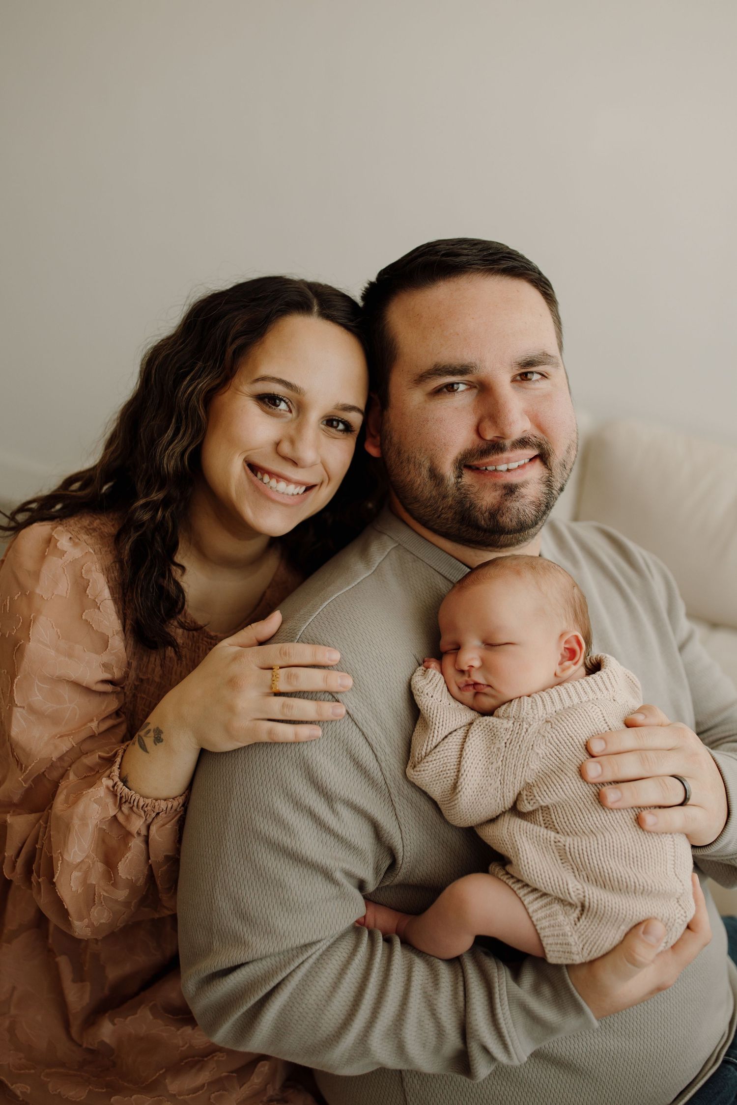 A couple in casual attire smiles while holding their sleeping newborn baby in a warm indoor portrait.