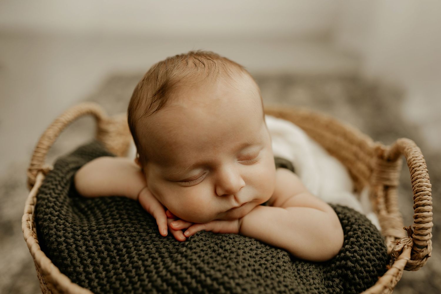 A sleeping newborn baby rests peacefully on a dark green knit blanket in a woven basket during a professional photoshoot.