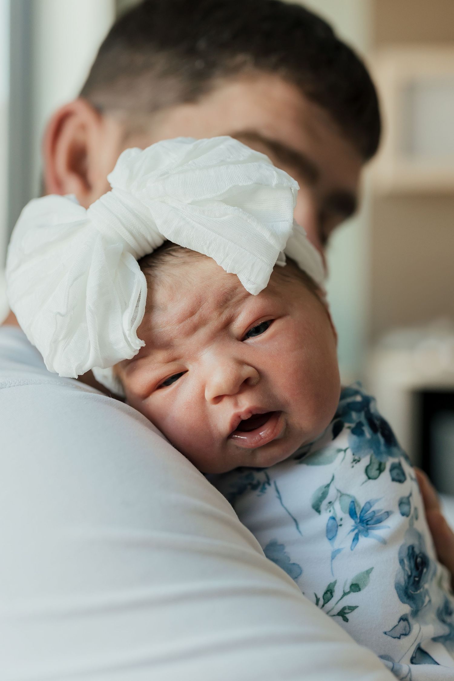 A newborn baby wearing a blue floral outfit rests peacefully against white bedding.