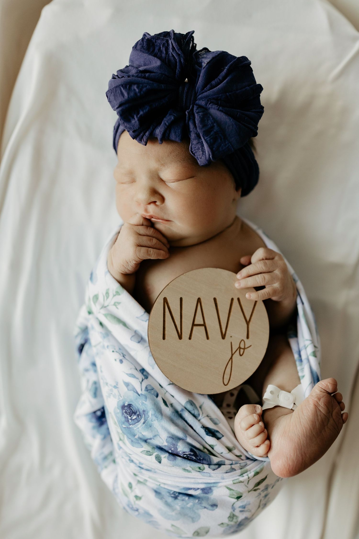 Newborn baby wrapped in floral swaddle with navy blue headband holding wooden name sign on white bedding.