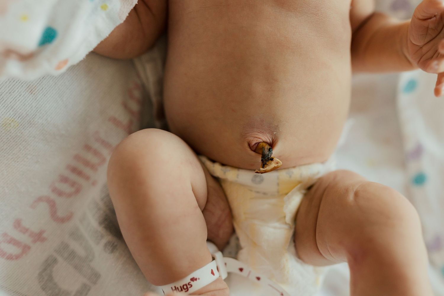 A small baby wearing a white diaper sits on a soft surface and plays with their toes.