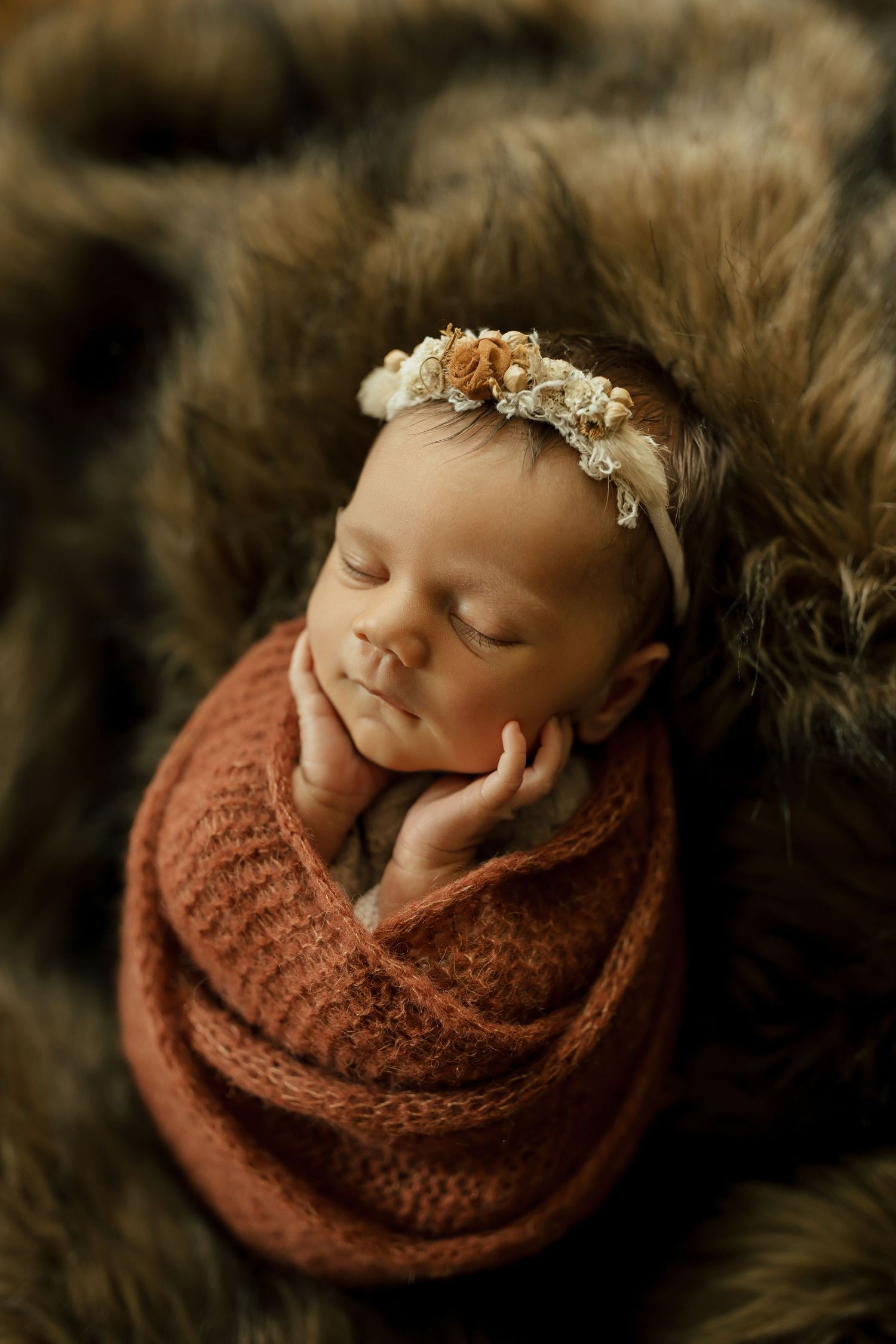 Newborn baby sleeping peacefully wrapped in brown knit blanket on soft fur backdrop with delicate floral headpiece.