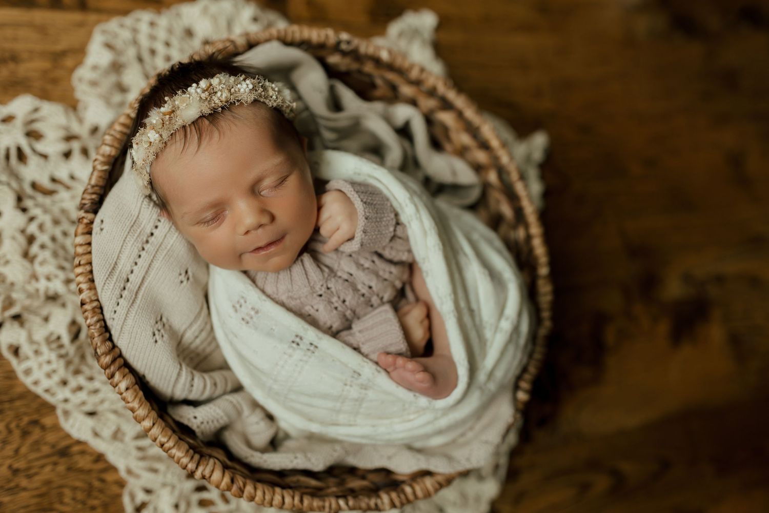 Newborn baby sleeping peacefully in a white blanket with delicate lace trim against dark brown background.