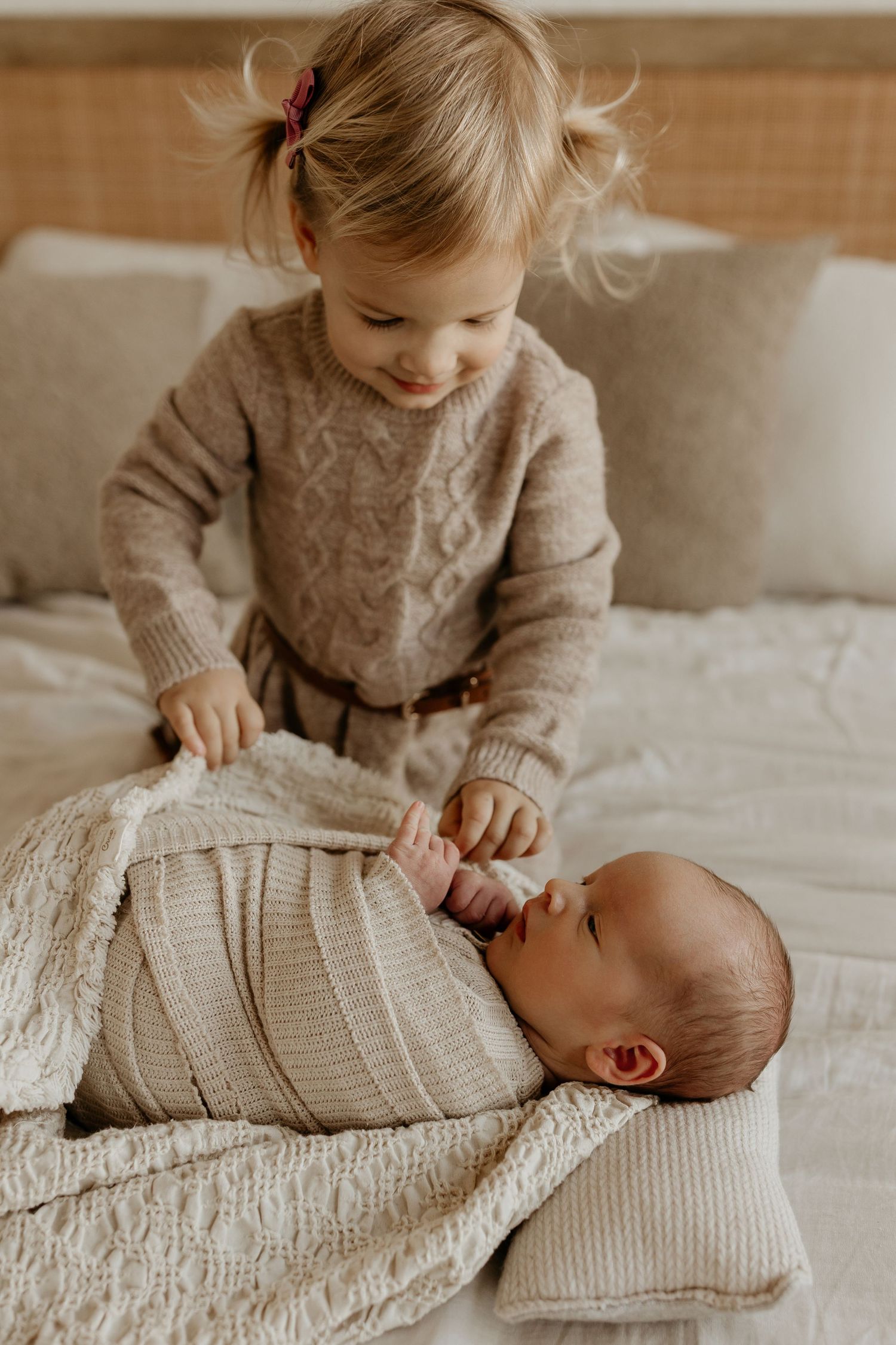 Siblings share a tender moment as older child in beige sweater gently interacts with baby in knit blanket on white bedding.