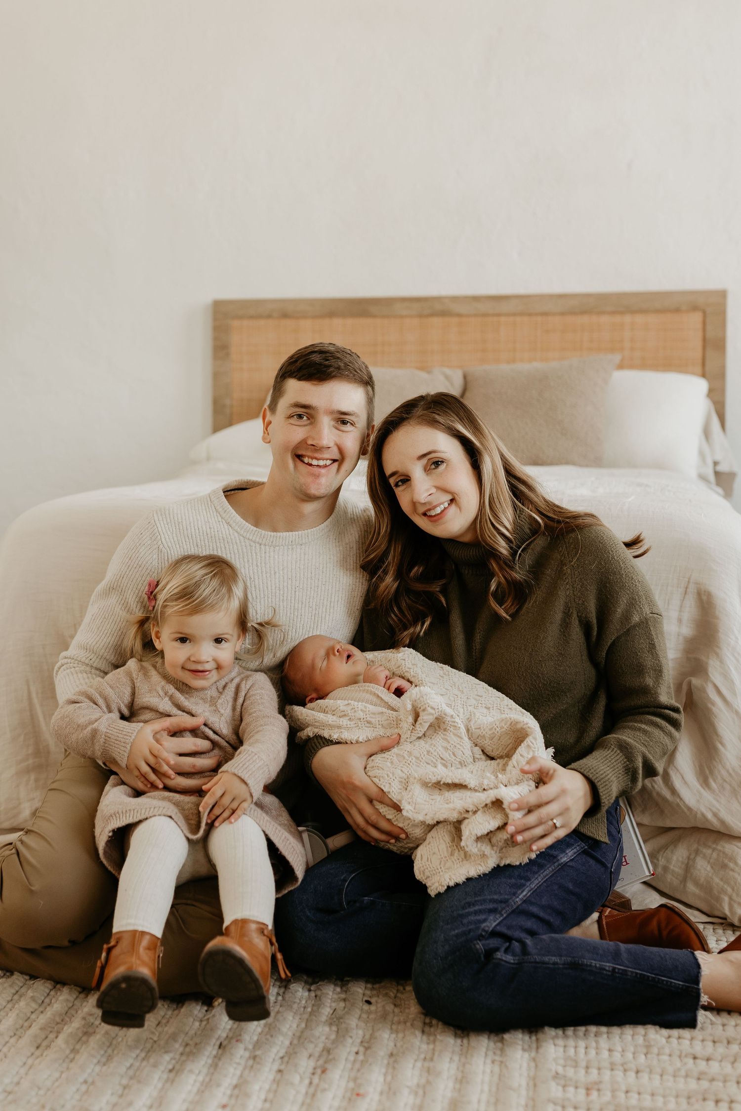 Family relaxes together on bed wearing cozy sweaters while bonding with their newborn in warm natural lighting.
