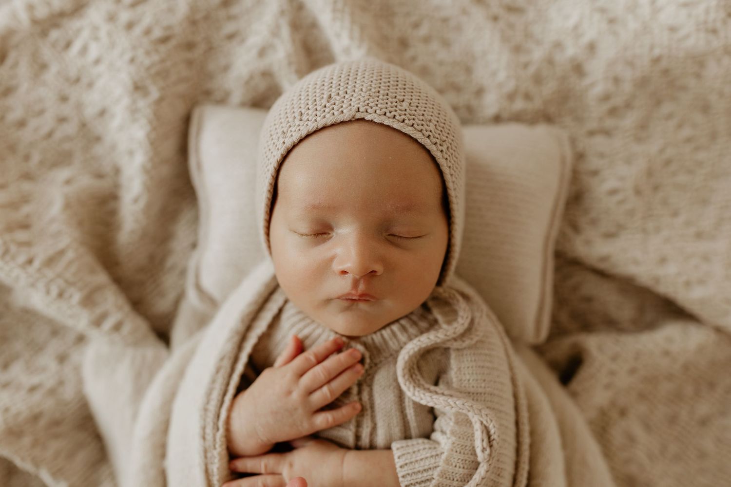 Sleeping baby wearing beige knit bonnet and sweater lies peacefully on white pillow with textured blanket.