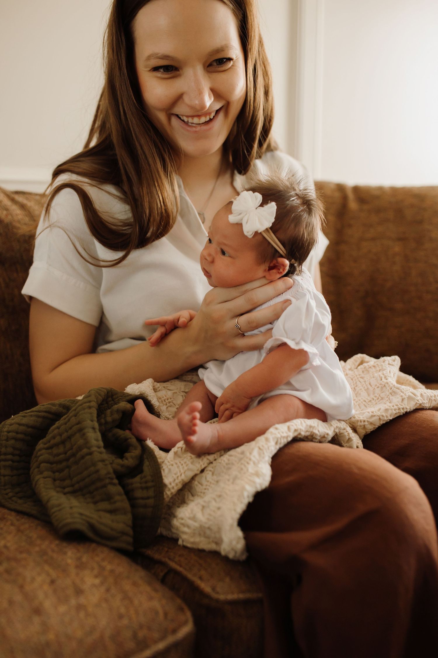 A tender moment between parent and baby in white dress sitting on brown couch in warm lighting.