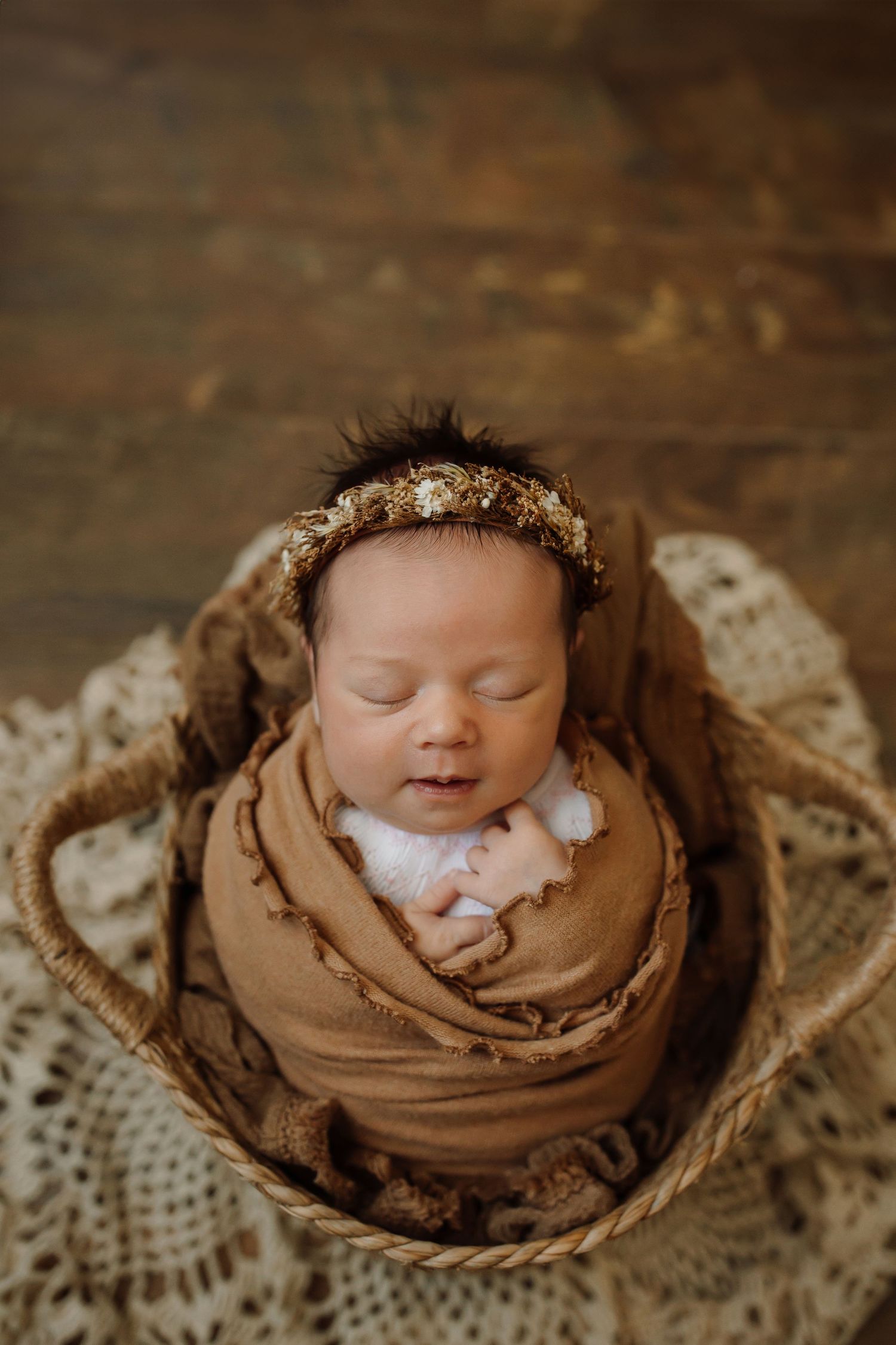 A sleeping newborn wrapped in brown fabric nestles peacefully in a rustic wooden setting.