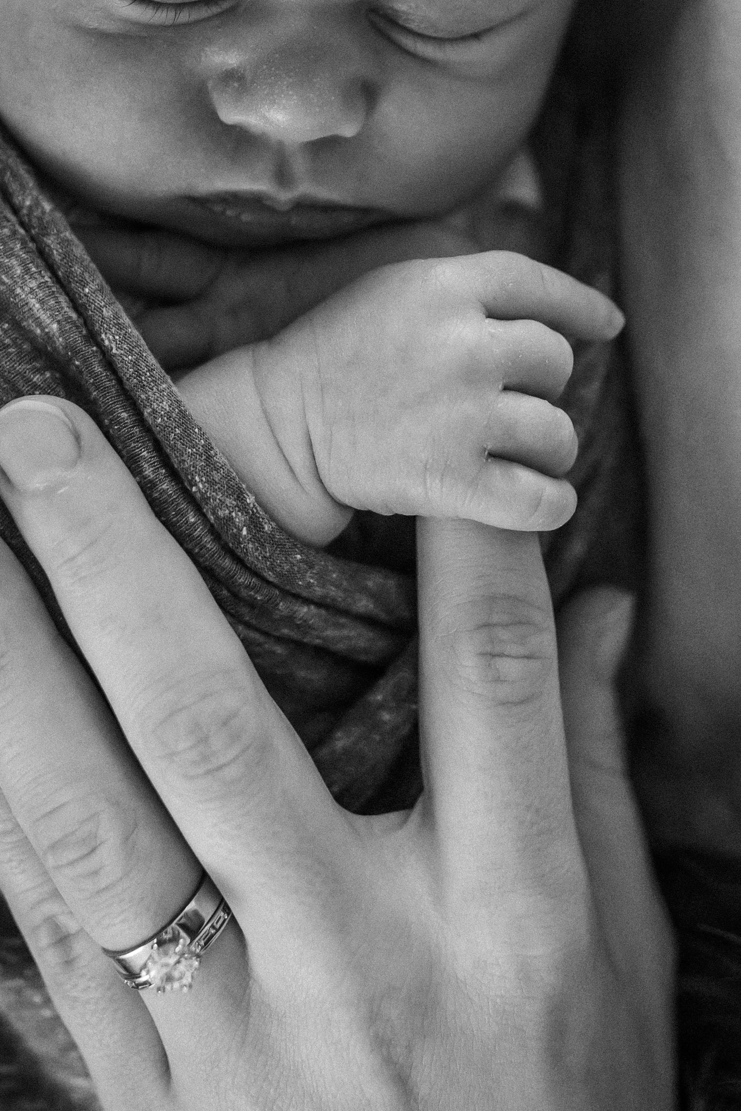 A tender black and white close-up of a baby's tiny hand grasping an adult finger wearing an engagement ring.