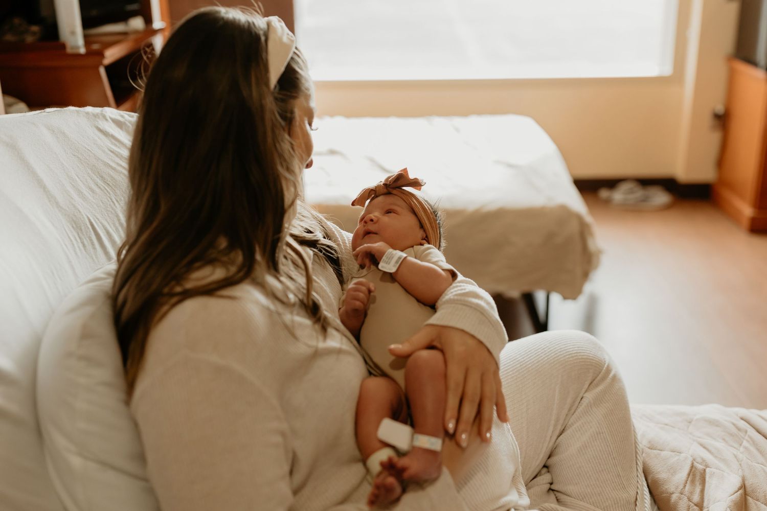 A person in a white sweater holds a small newborn baby while sitting on a bed in warm natural lighting.
