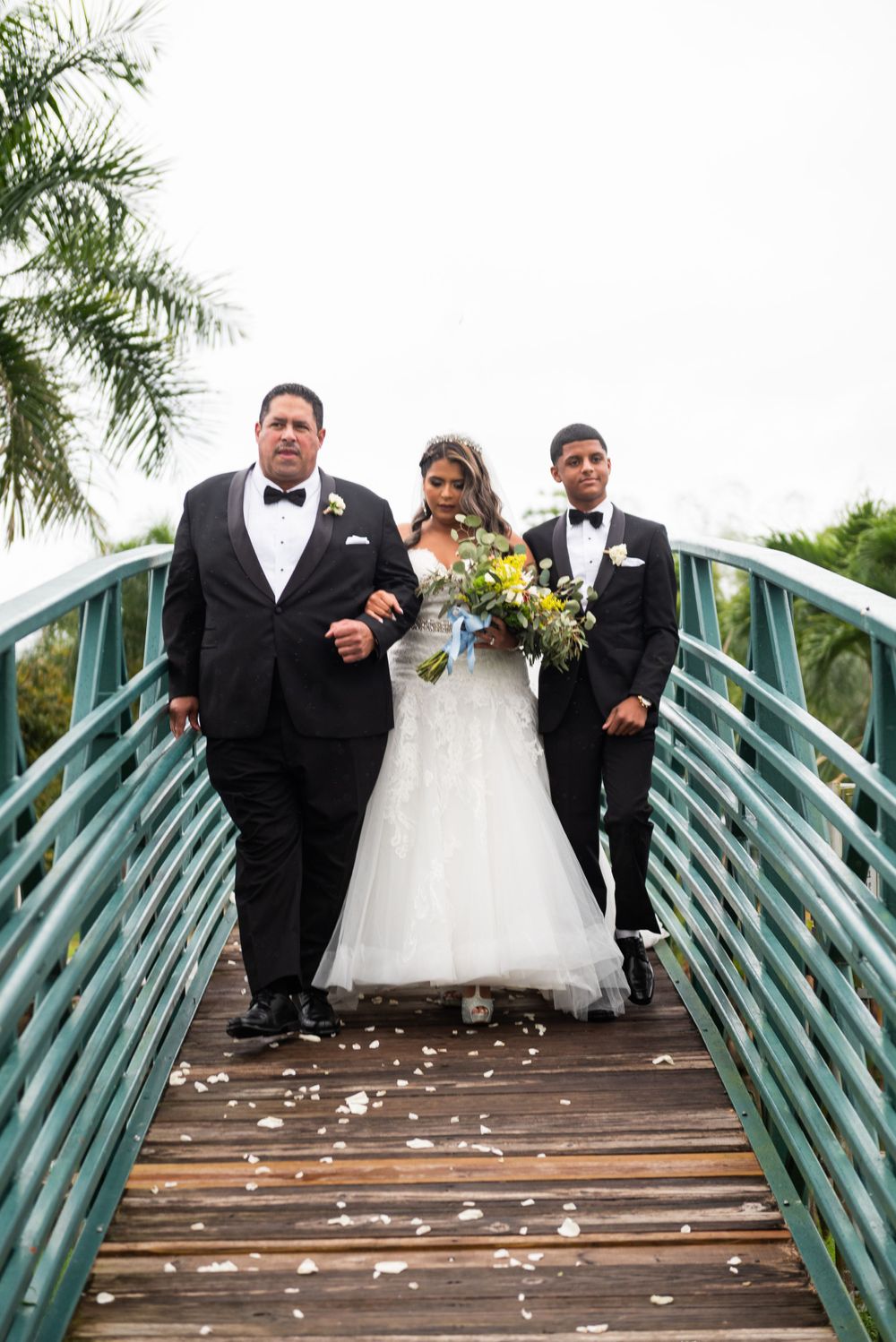 Wedding party walking across a teal-colored bridge with palm trees in the background.