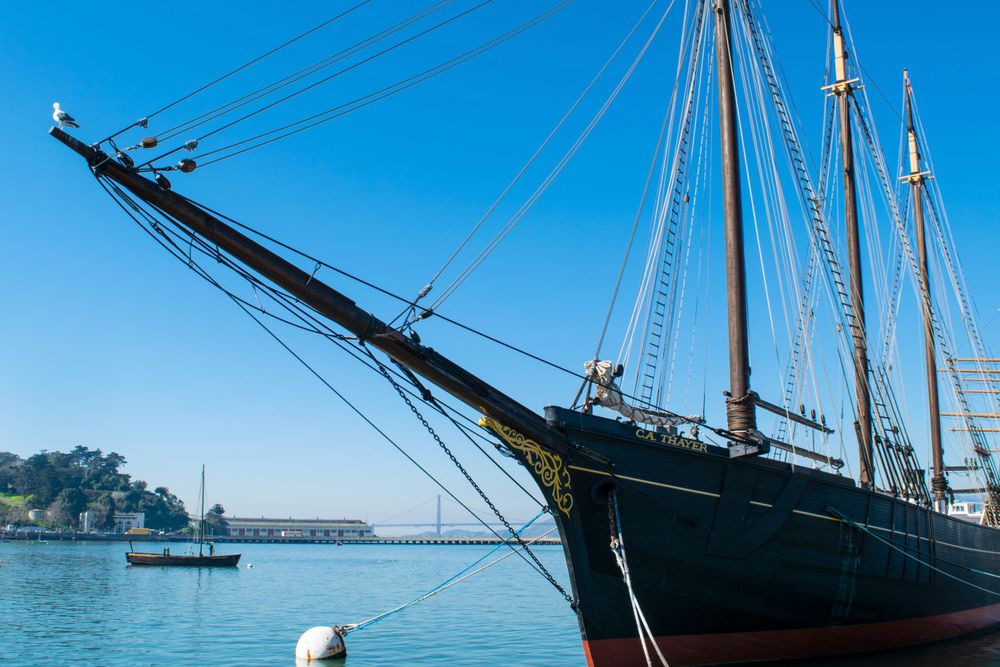 A historic tall ship with dark hull and multiple masts docked in a harbor with blue water.
