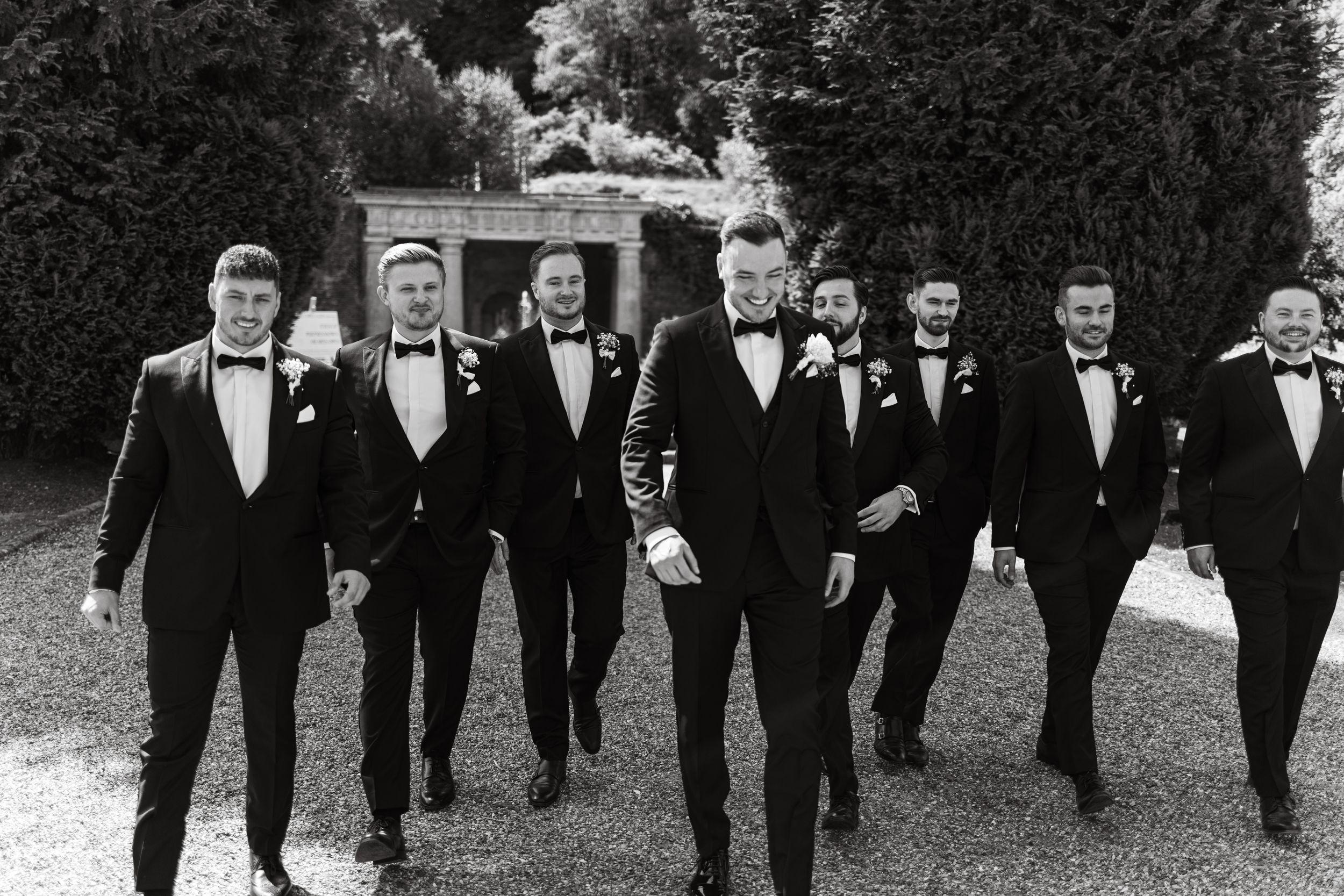 Groomsmen in black tuxedos walk together in formation outdoors.