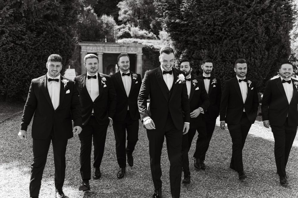 Groomsmen in black tuxedos walk together in formation outdoors.
