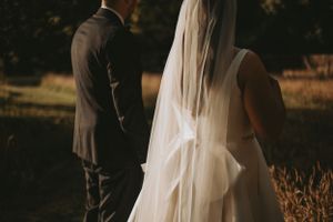 A wedding couple stands in a sunlit field with the bride's veil flowing in the breeze.