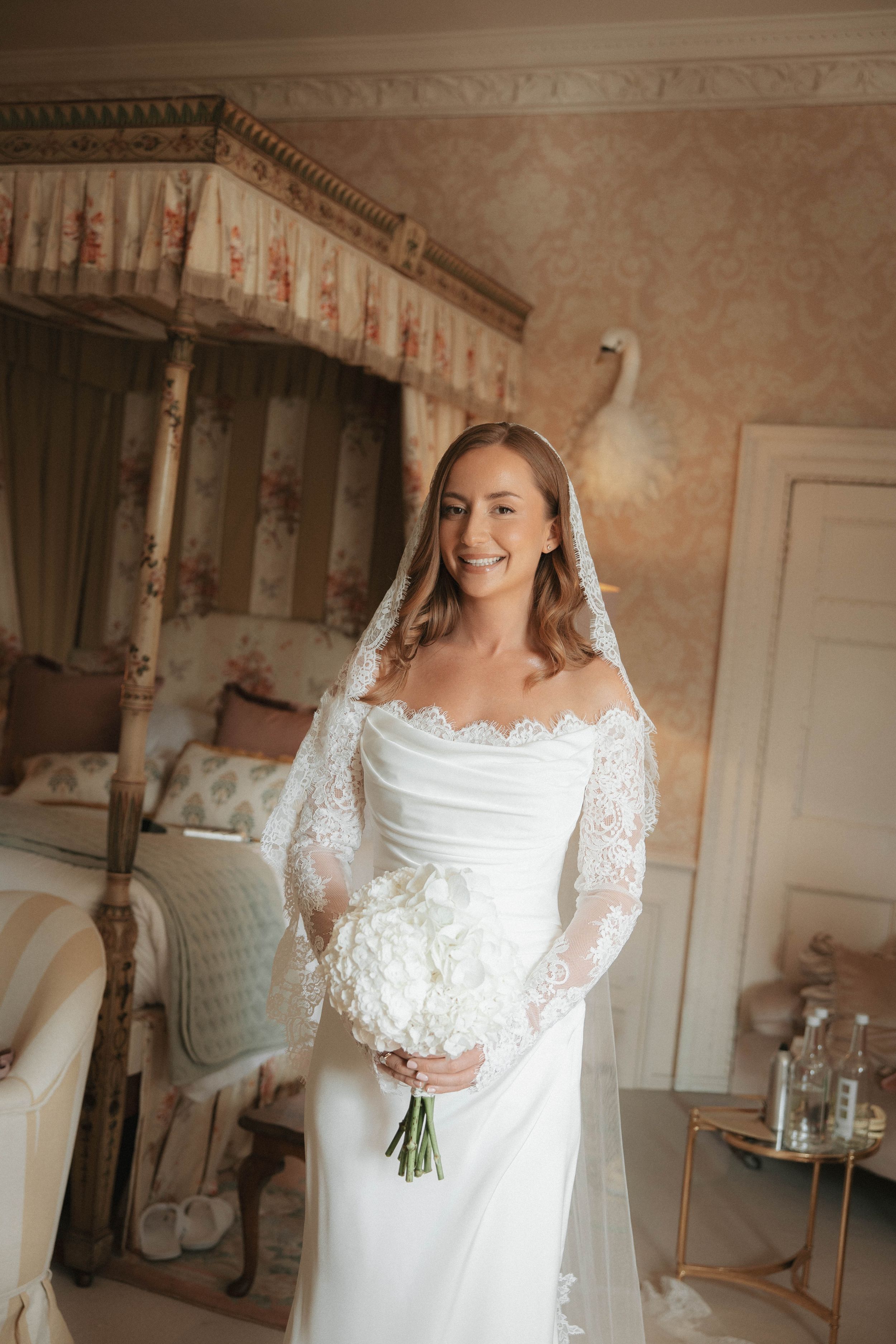 Smiling bride in lace veil and off-shoulder gown holds white bouquet in elegant floral bedroom.