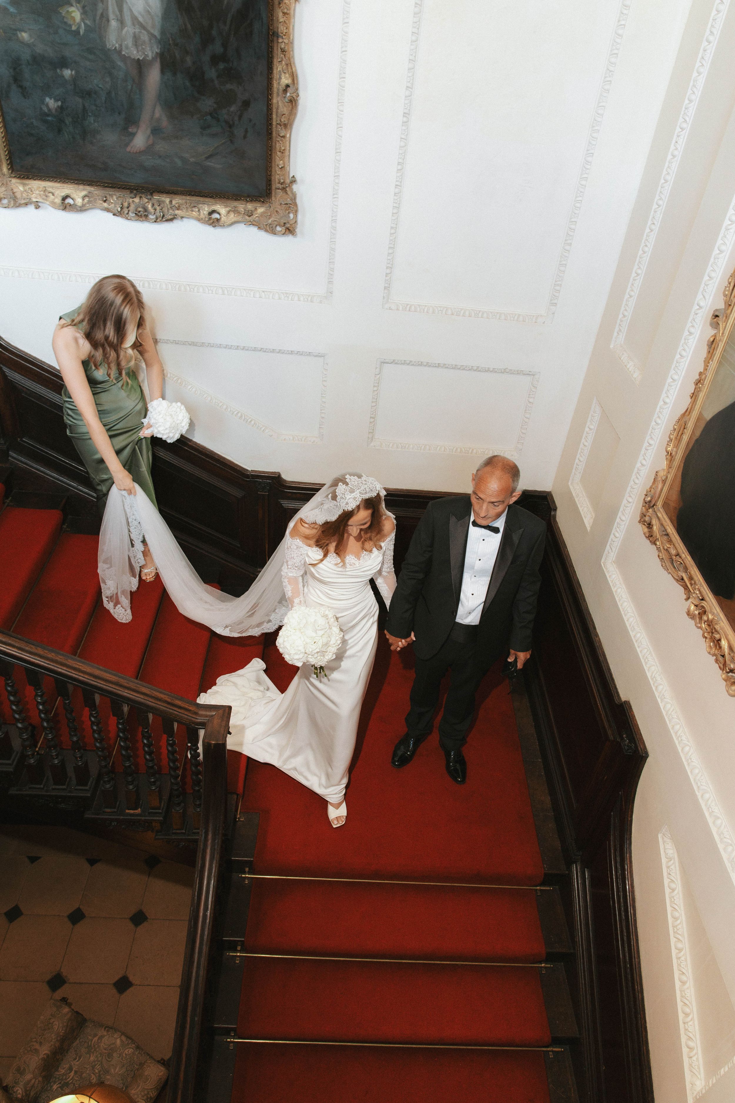 Bride in white gown walks down red-carpeted staircase with groom in tuxedo and bridesmaid carrying train.