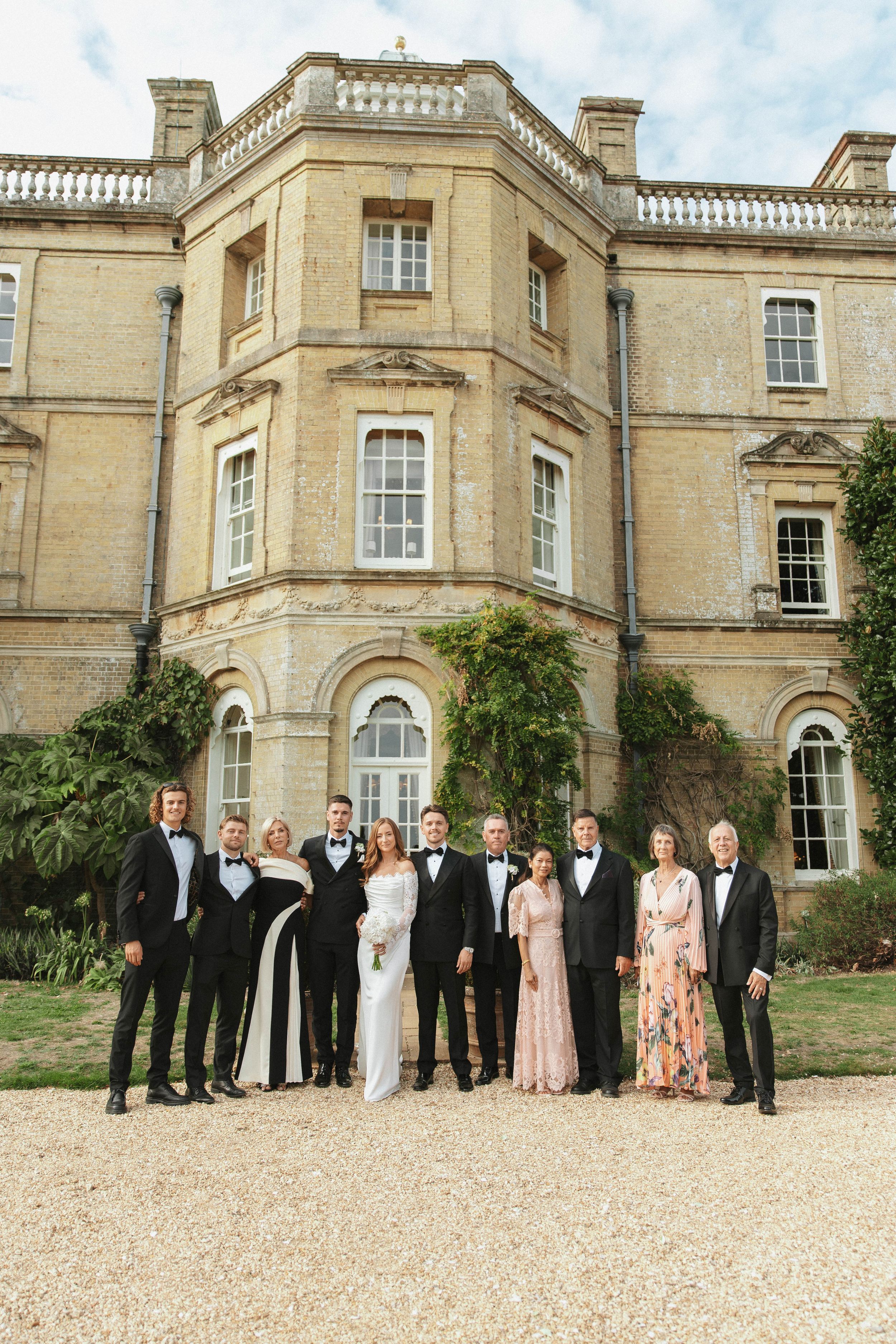 Wedding party posing in formal attire before a grand English manor house on a gravel path.