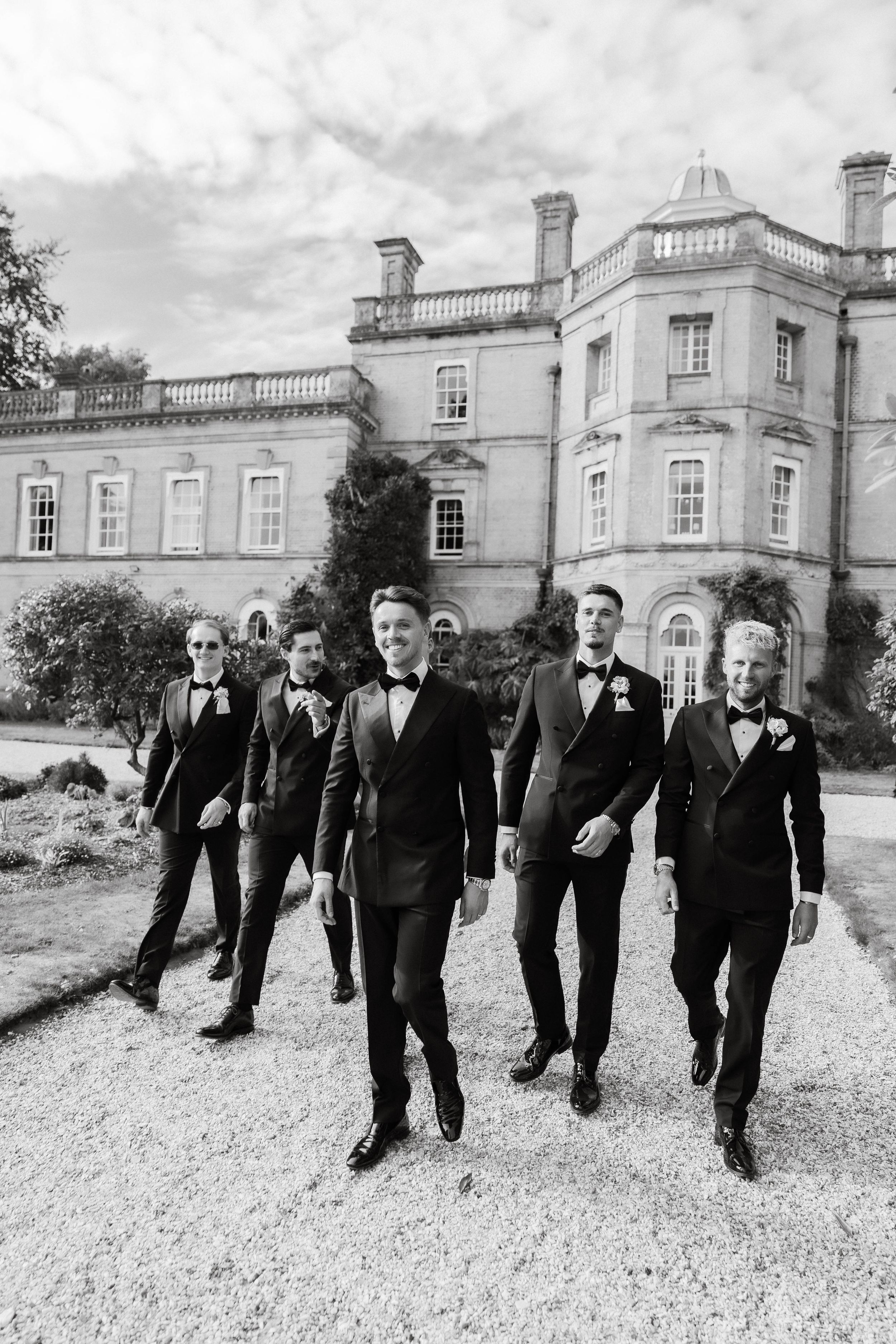 Groom and groomsmen in black tuxedos walk confidently in front of a grand estate in a black and white wedding photo.