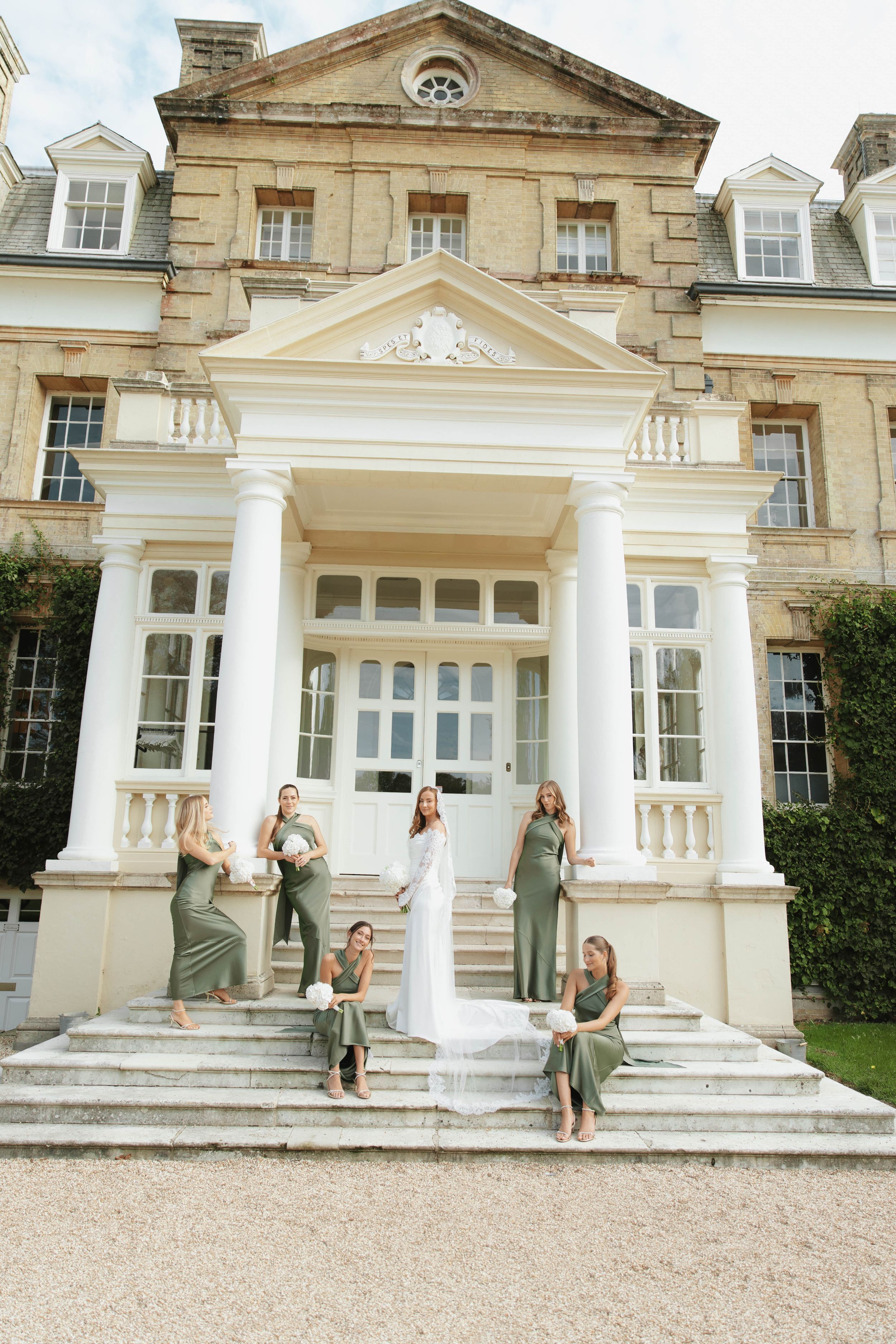 Bride in white gown poses with bridesmaids in sage green dresses on steps of elegant manor house.