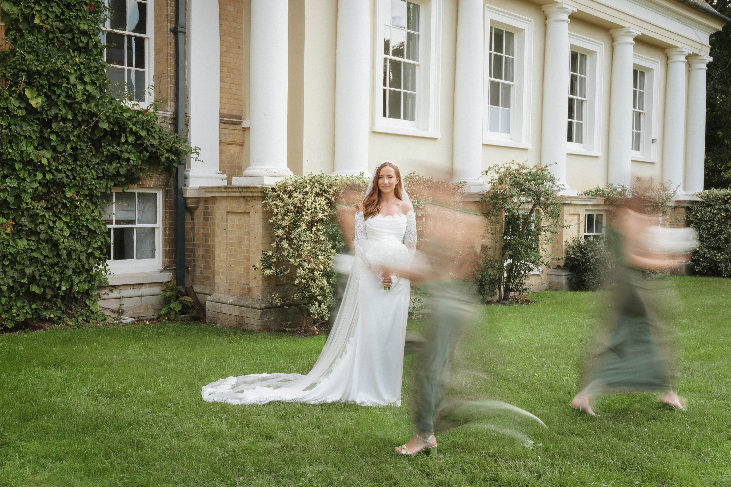 Pregnant woman in white lace gown poses outside elegant manor house with ivy walls while guests walk past.