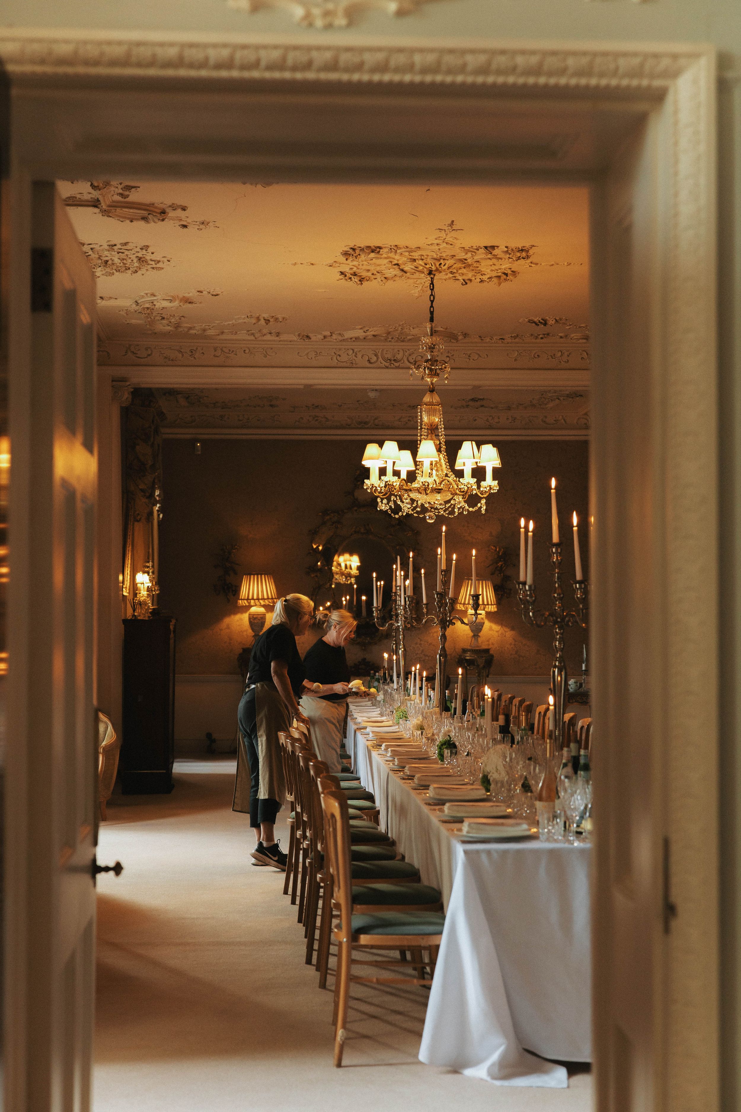 Elegant formal dining room with long white-clothed table, crystal chandelier, candelabras, and ornate ceiling moldings.