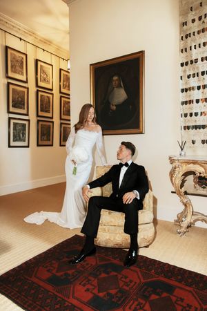 Bride in white gown stands beside groom in tuxedo seated in ornate chair in elegant room with portraits.