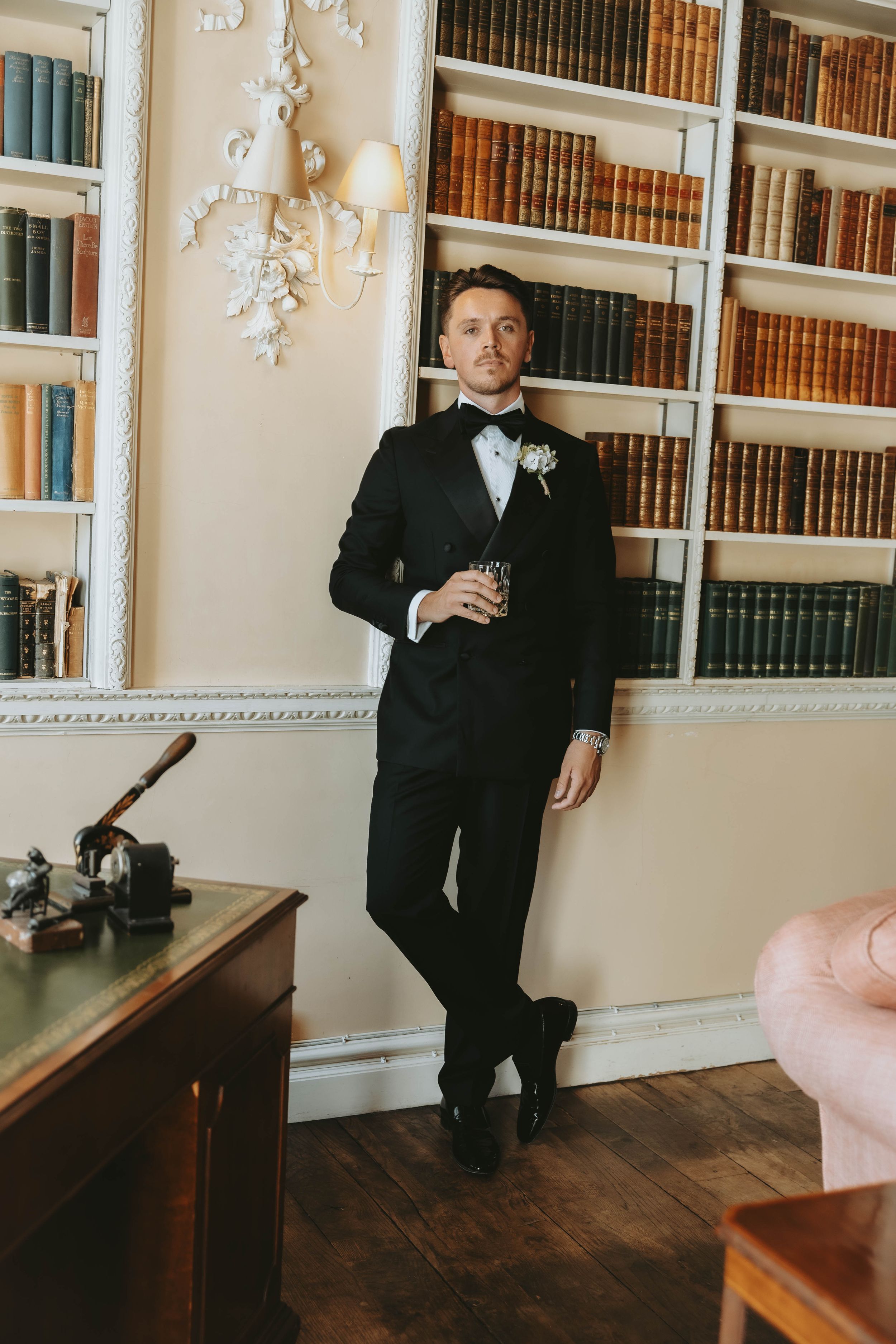 Groom in black tuxedo with boutonniere holds drink while leaning against bookshelf in elegant library.