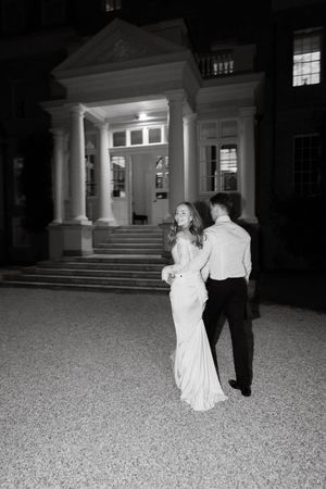Black and white photo of bride and groom walking toward an elegant illuminated mansion entrance at night.