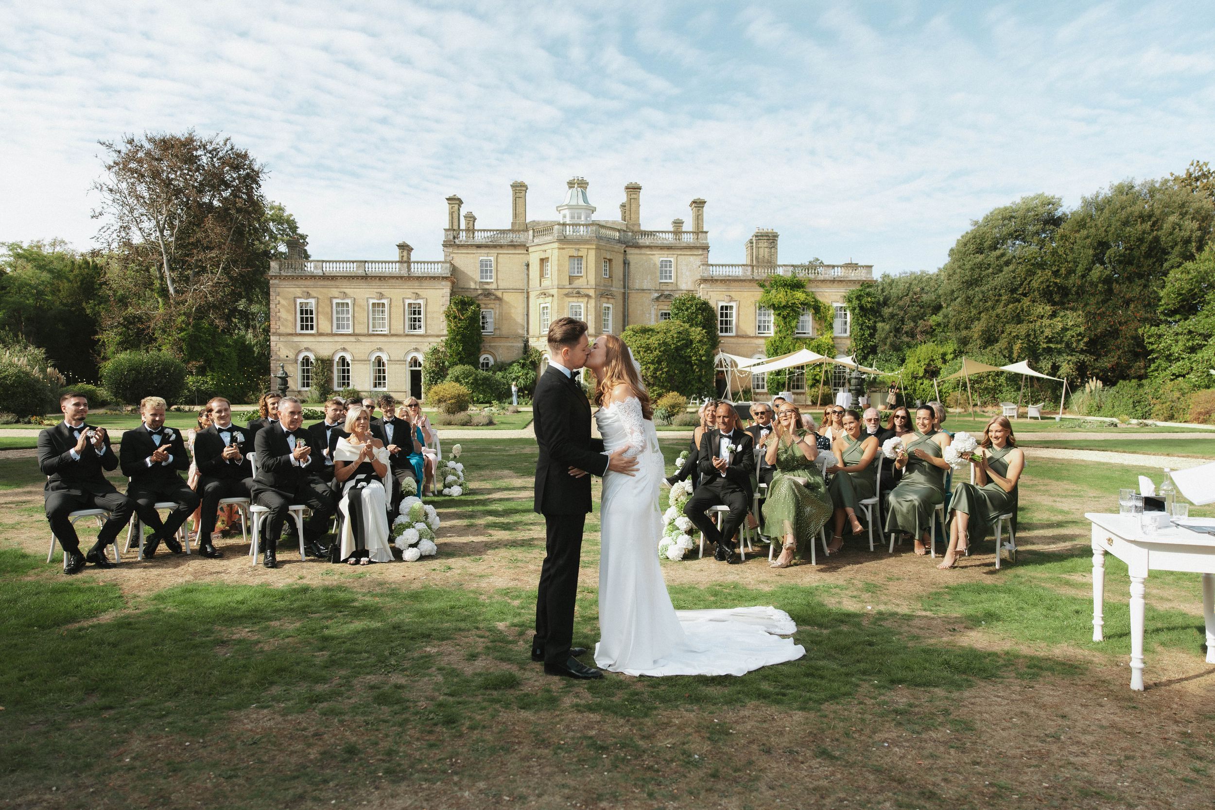 Outdoor wedding ceremony on manicured lawn before grand English manor house with seated guests.