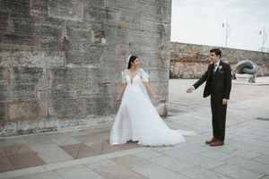 Bride in flowing white gown and groom in black suit pose against stone wall.