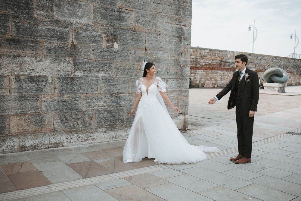 Bride in flowing white gown and groom in black suit pose against stone wall.