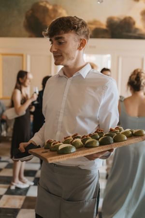 Server in white shirt carries wooden board with green macarons at wedding reception.