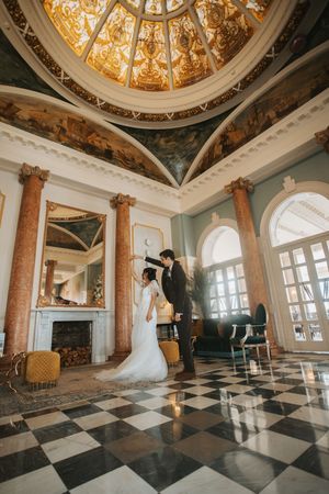 Elegant ballroom with ornate domed ceiling, marble columns and black and white checkered floor in panoramic view.