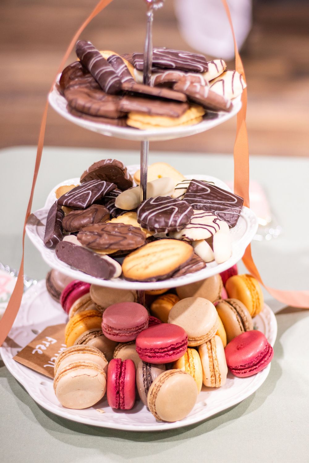Three-tiered dessert stand displaying chocolate-dipped cookies and colorful French macarons in pink, beige and brown shades.