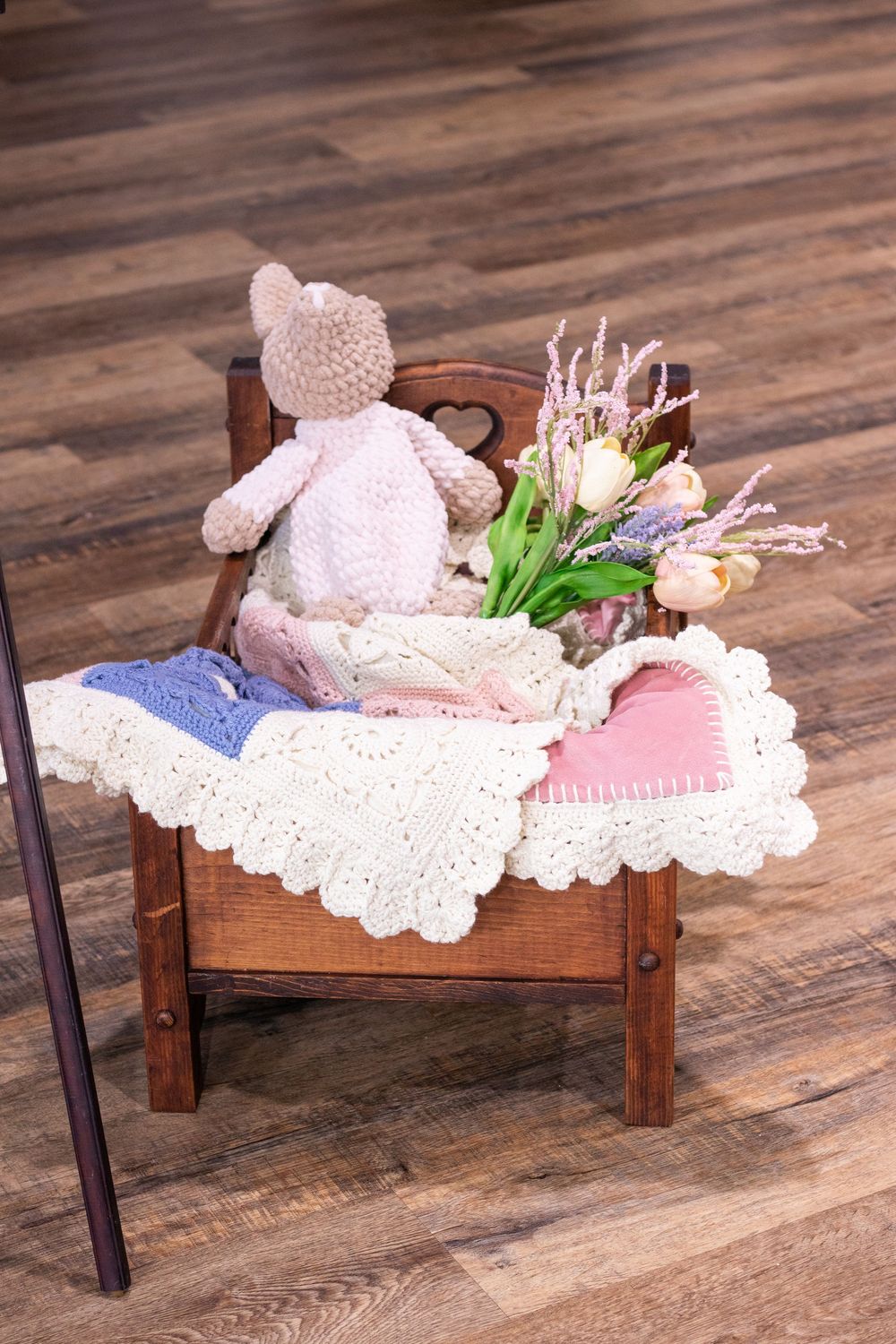 Pink crocheted teddy bear resting on white doily on wooden doll bed with decorative greenery.
